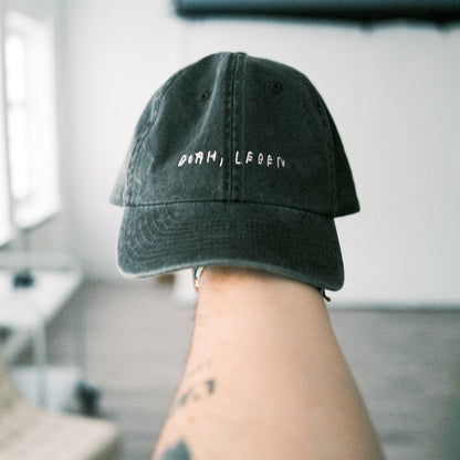 Schwarze Vintage-Cap aus Baumwollstoff mit verstellbarem Riemen hinten, gehalten von einer Hand im Vordergrund. Auf der Vorderseite der Cap steht in weißer, handschriftlich anmutender Schrift der Schriftzug „DORH, LEBEN“. Die Cap zeigt sieben kleine Lüftungsösen und eine gebogene Schirmkante.