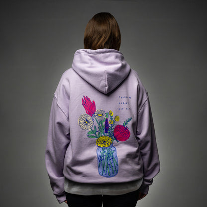 Rückansicht eines fliederfarbenen Hoodies, getragen von einer Person. Der Rücken zeigt einen großflächigen, farbenfrohen Blumenprint in einer Vase, daneben der handgeschriebene Text „perhaps nobody but you“.

