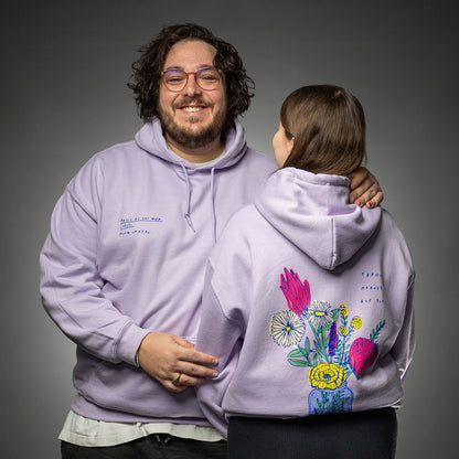 Zwei Personen tragen fliederfarbene Kapuzenpullover. Die Person links zeigt den Hoodie von vorn mit blauer Stickerei auf der Brust, die Person rechts präsentiert den Rückendruck: ein bunter Blumenstrauß in einer Vase, kombiniert mit dem englischen Text „perhaps nobody but you“.

