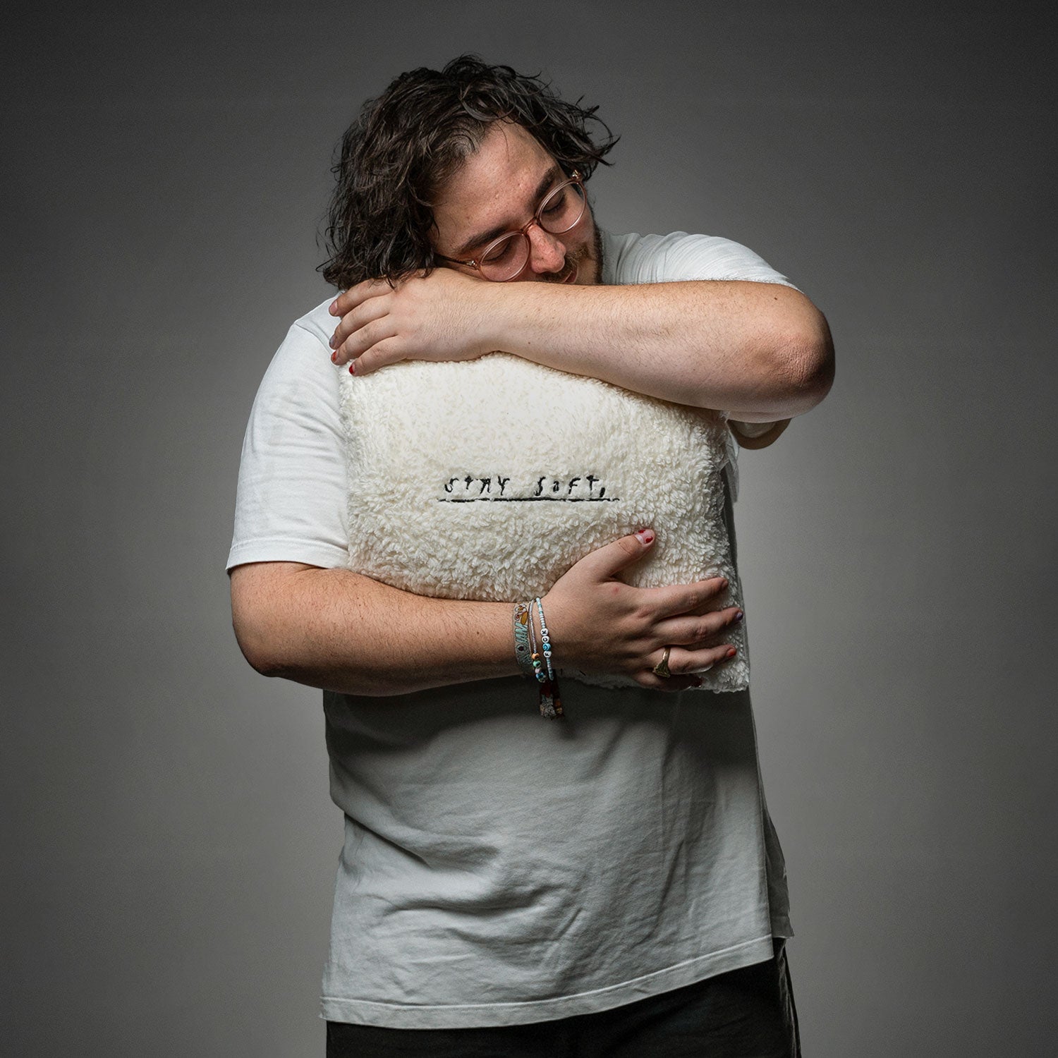 Eine Person im weißen T-Shirt hält ein quadratisches, flauschiges Kissen mit der gestickten Aufschrift „STAY SOFT,“ vor den Oberkörper, Gesicht nicht sichtbar.