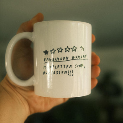 Weiße Keramiktasse mit schwarzer handschriftlicher Bewertung in Stern-Format auf der Vorderseite. Die Bewertung zeigt 1 von 5 Sternen. Darunter steht in handschriftlicher Schrift:
„FRAUEN WERDEN KOMPLETTER SCHAM, AUFPASSEN!!!“.
Die Tasse wird in einer Hand gehalten, unscharfer Hintergrund mit grünen Pflanzen.