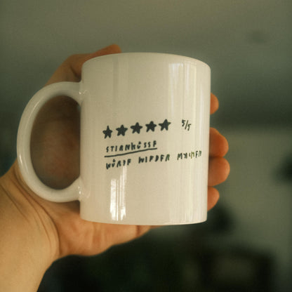 Weiße Keramiktasse mit schwarzer handschriftlicher Bewertung in Stern-Format auf der Vorderseite. Die Bewertung zeigt 5 von 5 Sternen. Darunter steht in handschriftlicher Schrift:
„STIRNKUSS – WÜRDE WIEDER NEHMEN“.
Die Tasse wird in einer Hand gehalten, unscharfer Hintergrund mit grünen Pflanzen.
