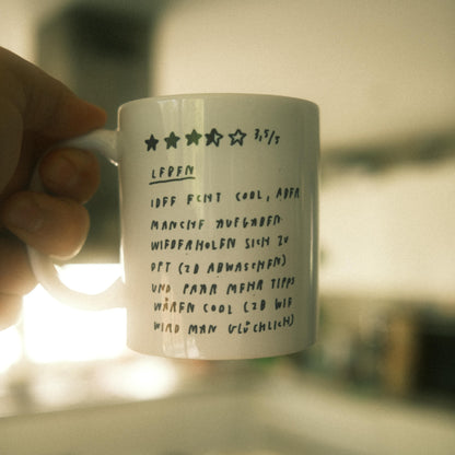 Weiße Keramiktasse mit schwarzer handschriftlicher Bewertung in Stern-Format auf der Vorderseite. Die Bewertung zeigt 3,5 von 5 Sternen. Darunter steht in handschriftlicher Schrift:
„LIEBEN
IDEE FINDET COOL, ABER MANCHMAL AUFGABEN WIEDERHOLEN SICH ZU OFT (ZB ABWASCHEN) UND PAAR MEHR TIPPS WÄREN COOL (ZB WIE WIRD MAN GLÜCKLICH)“.
Die Tasse wird von einer Hand gehalten, unscharfer Hintergrund.