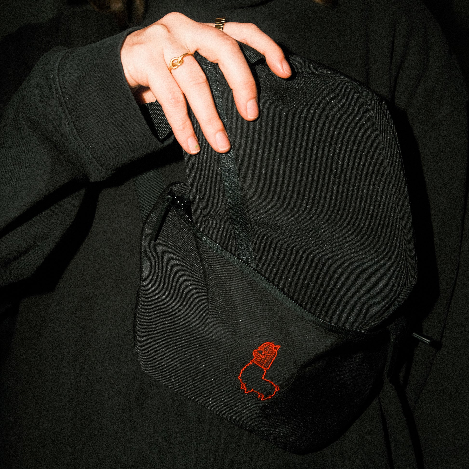 Schwarze Bauchtasche aus Stoff, die vor einem dunklen Oberteil gehalten wird. Eine Hand mit goldenen Ringen öffnet den oberen Reißverschluss der Tasche. Auf der Vorderseite ist eine kleine, rot gestickte Alpaka-Kontur mit Mütze zu sehen. Die Szene ist dunkel gehalten, wodurch Tasche und Stickerei kontrastreich hervortreten.