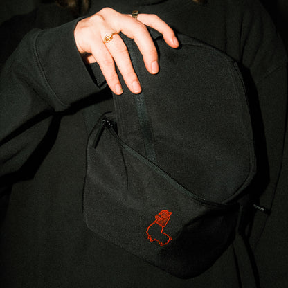 Schwarze Bauchtasche aus Stoff, die vor einem dunklen Oberteil gehalten wird. Eine Hand mit goldenen Ringen öffnet den oberen Reißverschluss der Tasche. Auf der Vorderseite ist eine kleine, rot gestickte Alpaka-Kontur mit Mütze zu sehen. Die Szene ist dunkel gehalten, wodurch Tasche und Stickerei kontrastreich hervortreten.