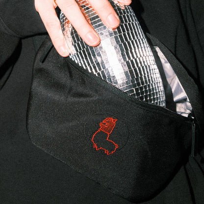 Schwarze Bauchtasche mit roter Alpaka-Stickerei auf der Vorderseite. Eine Hand zieht eine glänzende Discokugel-Flasche aus dem geöffneten Reißverschlussfach. Die Tasche wird vor einem dunklen Oberteil getragen, die Szene ist stark ausgeleuchtet und kontrastreich inszeniert.