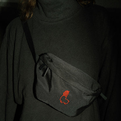 Schwarze Bauchtasche aus Stoff, quer über einen dunklen Rollkragenpullover getragen. Auf der Vorderseite der Tasche ist eine kleine, rot gestickte Alpaka-Kontur mit Mütze zu sehen. Die Szene ist schwach beleuchtet, wodurch Tasche und Stickerei besonders hervorgehoben werden.