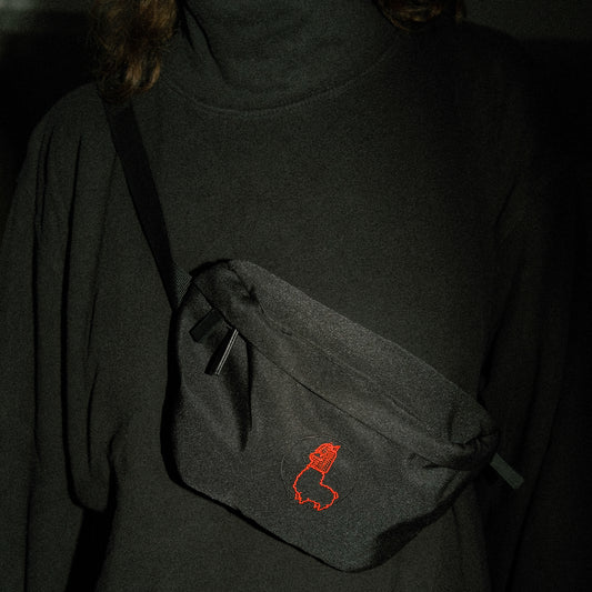 Schwarze Bauchtasche aus Stoff, quer über einen dunklen Rollkragenpullover getragen. Auf der Vorderseite der Tasche ist eine kleine, rot gestickte Alpaka-Kontur mit Mütze zu sehen. Die Szene ist schwach beleuchtet, wodurch Tasche und Stickerei besonders hervorgehoben werden.