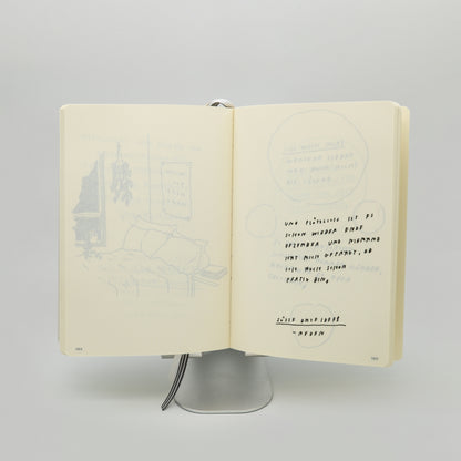 Aufgeschlagenes Buch auf silbernem Ständer. Links eine blaue Zeichnung eines Schlafzimmers mit Bett, Fenster und Pflanzen. Rechts handgeschriebener Text in schwarzer Schrift:
„Und plötzlich ist es schon wieder Ende Dezember und niemand hat mich gefragt, ob ich auch schon fertig bin,“
darunter unterstrichen:
„Süße Dateidee – Reden“.
Zwei Lesezeichenbänder hängen unten heraus.