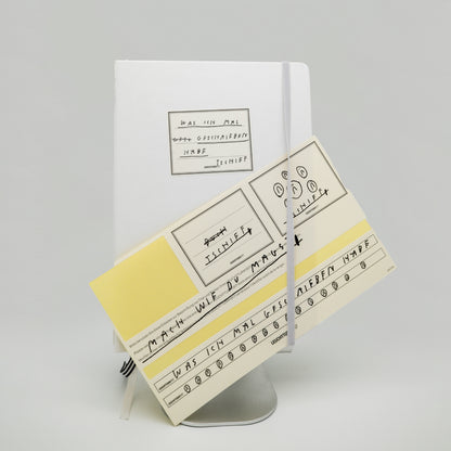Weißes Buch mit Gummiband auf silbernem Ständer. Auf dem Buchdeckel ein handgeschriebener Aufkleber mit Text:
„Was ich mal
esse geschrieben
habe
Tschiff“.
Vor dem Buch befinden sich gelbe und weiße Aufkleberbögen mit handgeschriebenen Texten und kleinen Gesichtern.
Am unteren Rand eines Aufklebers steht:
„Was ich mal geschrieben habe Tschiff“.