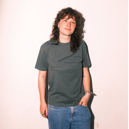 Dunkelgrünes T-Shirt mit kurzen Ärmeln. Auf der Brust steht in schwarzer Handschrift der Text:

„FALLS DU DAS HIER LIEST: NOCH IMMER“