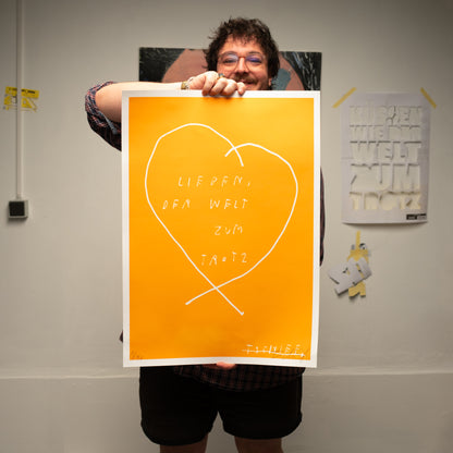 Person hält ein leuchtend orangefarbenes Poster vor sich, das fast den gesamten Oberkörper verdeckt. In der Mitte des Posters ist ein großes, handgezeichnetes Herz mit weißer Linie zu sehen. Innerhalb des Herzens steht in weißer, handgeschriebener Schrift der Text: „LIEBEN, DER WELT ZUM TROTZ“. Unten rechts ist eine Signatur zu erkennen, unten links eine handschriftliche Editionsnummer. Im Hintergrund hängt an der Wand ein weiteres Poster mit grün-weißem Textmotiv.