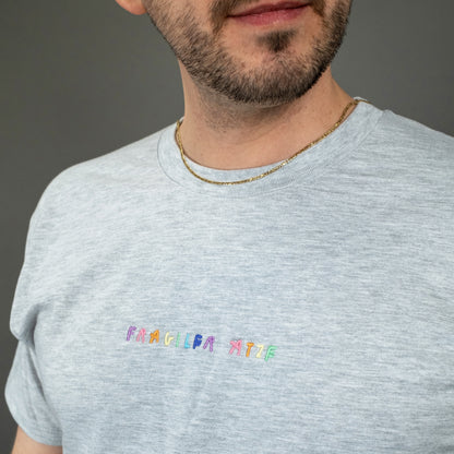 Nahaufnahme eines hellgrauen T-Shirts mit kurzärmeligem Schnitt. Auf der linken Brust ist mittig eine bunte Stickerei mit dem Schriftzug „FRAGILER ATZE“ in verschiedenen Farben zu sehen. Der Stoff wirkt weich und leicht meliert.