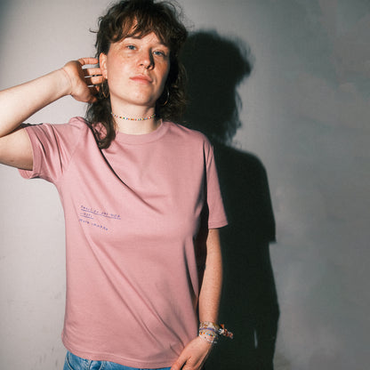 Altrosa T-Shirt mit kleiner, handgeschriebener Textstickerei auf der linken Brustseite. Der gestickte Text in Dunkelblau lautet: „Sag es der Welt: Liebe ist radikal“. Das T-Shirt wird von einer Person getragen, die locker eine Hand in den Nacken legt. Sie trägt bunte Accessoires wie ein Perlenarmband und ein Perlenhalsband. Der Hintergrund ist neutral, mit starker seitlicher Lichtführung und deutlichem Schlagschatten.