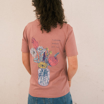 Altrosa T-Shirt mit großem Rückendruck. Der Druck zeigt ein gezeichnetes Bouquet aus verschiedenen Blumen in einer transparenten Vase. Die Blumen sind farbenfroh illustriert in Gelb, Weiß, Rosa, Rot und Blau. Über dem Blumenmotiv steht in handgeschriebener blauer Schrift: „PERHAPS NOBODY BUT YOU.“ Das T-Shirt wird von einer Person getragen, die mit dem Rücken zur Kamera steht. Sie hat lockiges, schulterlanges Haar und trägt eine locker sitzende, hellblaue Jeans. Der Hintergrund ist schlicht und hell.