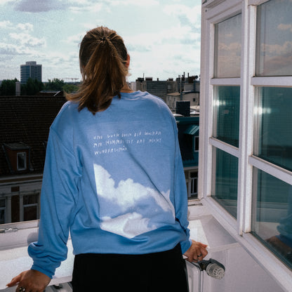Frau mit braunem Pferdeschwanz steht mit dem Rücken zur Kamera auf einem Balkon. Sie trägt ein hellblaues Sweatshirt mit weißem Wolkenmotiv und weißem handschriftlichem Text auf dem Rücken: „Und guck doch die Wolken am Himmel ist das nicht wunderschön“. Die Frau lehnt sich mit beiden Händen auf das Balkongeländer. Im Hintergrund sind Dächer und Gebäude einer Stadt zu sehen unter einem wolkigen Himmel.
