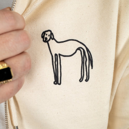 Detailaufnahme eines beigen Kapuzenpullovers mit Reißverschluss. Im Fokus steht ein handgezeichneter Hund in schwarzer Farbe auf der linken Brustseite des Pullovers. Die Zeichnung ist einfach und skizzenhaft gehalten.