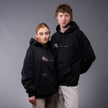 Zwei Personen tragen schwarze Kapuzenpullover mit rosa Stickdetails. Die Hoodies zeigen eine florale Liniengrafik mit Rose auf Brusthöhe; auf dem linken Hoodie ist zusätzlich der gestickte Schriftzug „Ich wünschte, die Welt wäre weicher“ über der Tasche zu sehen. Beide stehen nebeneinander vor neutralem, grauem Hintergrund.