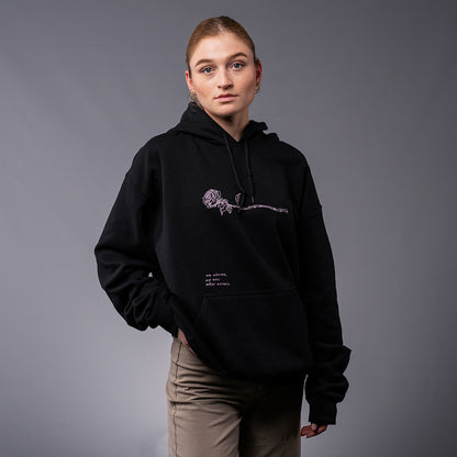 Model trägt den schwarzen Hoodie mit floraler Stickerei auf der Brust und dem Schriftzug „Ich wünschte, die Welt wäre weicher“ über der Bauchtasche. Die Kapuze ist aufgesetzt, das Outfit wird mit beiger Hose kombiniert.