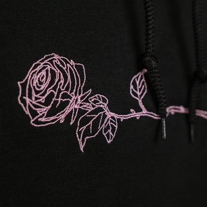 Detailansicht des Brustbereichs des schwarzen Hoodies mit einer floralen Stickerei. Die rosa Liniengrafik zeigt eine liegende Rose mit Blättern entlang der Brustpartie.

