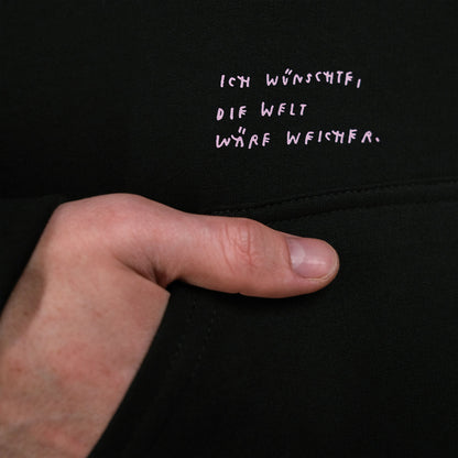 Nahaufnahme des schwarzen Hoodies mit rosa Schriftzug über der Kängurutasche. Der gestickte Text lautet: „Ich wünschte, die Welt wäre weicher.“
